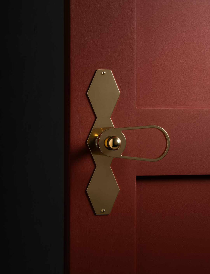 Poignée de porte goutte ajourée avec bouton bille, plaque festonnée géométrique en laiton naturel, mise en situation sur porte bordeaux à moulures – collection Feston