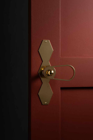 Poignée de porte goutte ajourée avec bouton bille, plaque festonnée géométrique en laiton naturel, mise en situation sur porte bordeaux à moulures – collection Feston